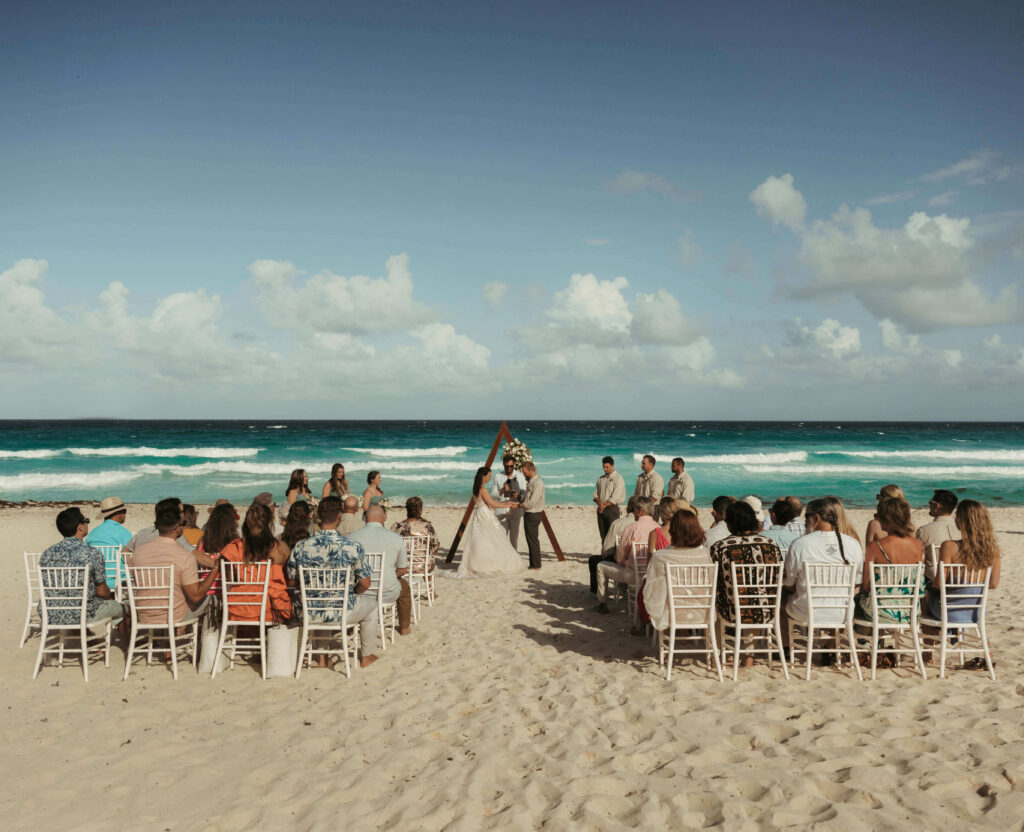 Bride and groom exchanging vows under a wooden triangle arch on the beach with guests seated in white chairs at Live Aqua Cancun
