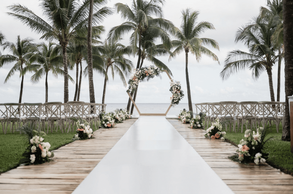Oceanfront ceremony setup with a floral hexagon arch, white aisle runner and palm trees at Grand Velas